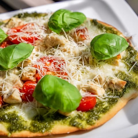 Freshly baked Chicken Pesto Pizza on a white ceramic platter, topped with tender shredded chicken, melted cheese, and bright green basil leaves.  